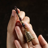 A close-up of a hand holding a brass bell keychain with a dark brown cord, decorative beads, and a small hanging bell against a dark background.