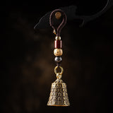 A hanging decorative bell ornament with a dark brown braided cord, gold-toned metal bell, and several accent beads in red, gold, and black, shown against a dark studio background.