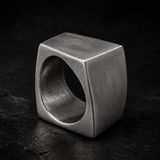 A chunky, geometric cube-shaped ring made of brushed silver-toned metal, photographed on a dark textured surface.
