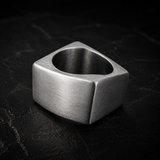 A brushed silver metal cube ring with a circular center hole, resting on a dark textured stone surface.