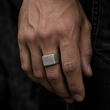 A close-up of a man's hand wearing a wide, rectangular silver-toned metal ring with a matte finish, set against a dark background.