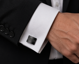 A close-up of a black textured cufflink with a woven pattern, attached to a white dress shirt cuff on a man's wrist, with a black suit jacket visible.