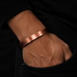 A minimalist copper-toned metal bangle bracelet worn on a person's wrist, shown in a close-up shot against a dark black background.