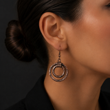A close-up of a model's ear wearing a textured bronze double-ring dangle earring against a dark background.