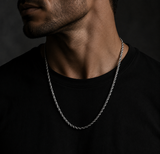 A close-up of a man wearing a silver twisted rope chain necklace over a black t-shirt against a dark background.