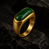 A gold signet-style ring with a large oval green stone, photographed resting on dark textured rock against a dark background.