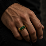 A close-up of a man's hand wearing a gold signet ring with a rectangular green stone, set against a dark background.