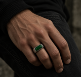 A close-up of a man's hand wearing a silver ring with a rectangular green stone, resting on his thigh.