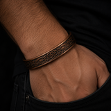 A close-up of a person's wrist and hand wearing a thick, engraved bronze-toned cuff bracelet with an ornate scroll pattern, set against a dark shirt and jeans background.