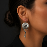 A close-up of a woman's ear wearing a silver dangle earring with a circular patterned top and dangling geometric elements, set against a dark background.