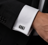 A close-up of a man's wrist wearing a silver-toned metal cufflink with three black stripes, paired with a white dress shirt and a black suit jacket.