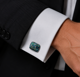 A close-up of a man's wrist wearing a black suit jacket and white dress shirt cuff, featuring a rectangular silver-toned cufflink with an intricate turquoise, green, and blue mosaic inlay.