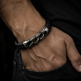 A close-up of a man's hand resting in his jeans pocket, wearing a silver-toned metal chain bracelet with engraved details and a black braided leather accent.