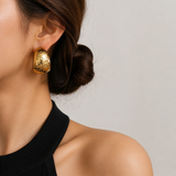 A close-up of a model's ear wearing a chunky, hammered gold hoop earring, with her hair in a low bun and a black ribbed top against a neutral light gray background.