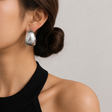 A close-up of a woman's ear wearing a hammered silver hoop earring, with her hair in a low bun and a black ribbed top against a light gray background.